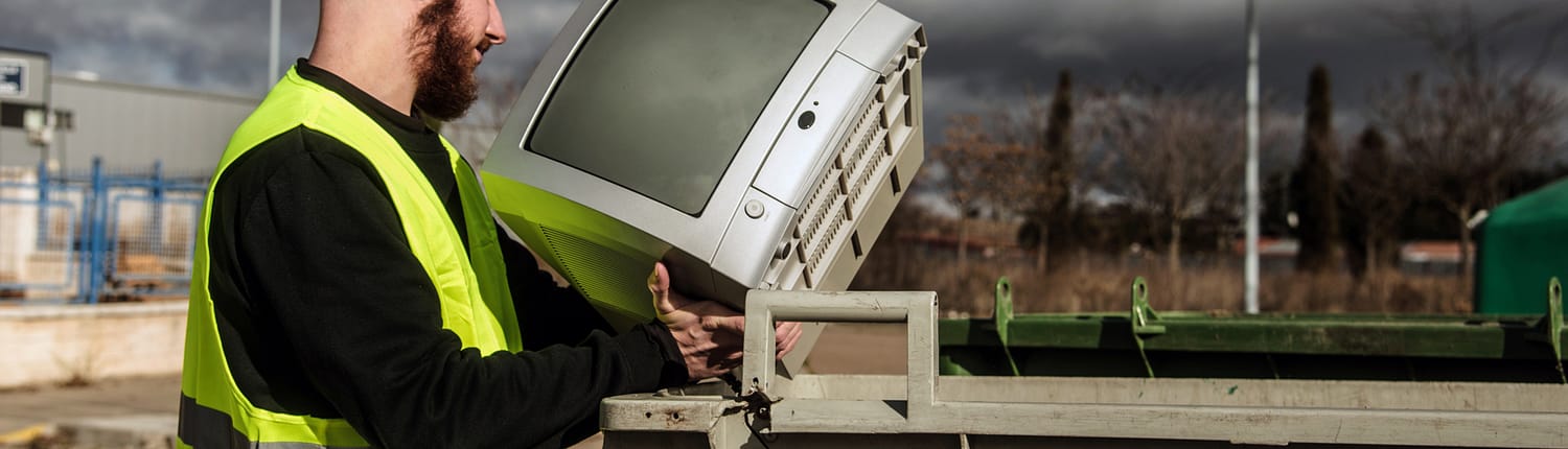 Recycling center worker throwing an old tube television into a grey dumpster.